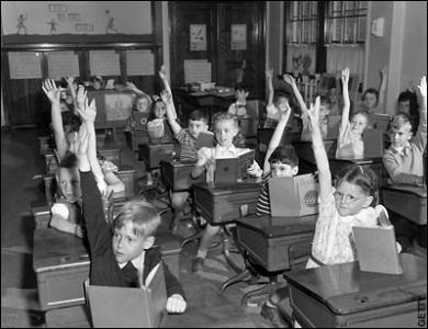 Old photo of young students in a classroom, everyone's hand raised to answer a question.